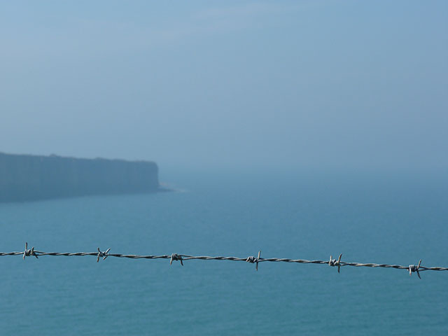 Photo d'un bord de mer sous la brume, sur le devant de l'image un fil barbelé